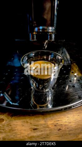 Modern design glass cup of glass with freshly brewed black coffee with golden foam with small bubbles on top of an automatic barista espresso machine Stock Photo