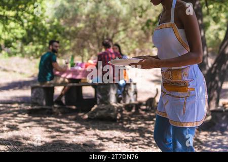 African American woman serving grilled food to friends at a forest picnic, with selective focus on the foreground. Stock Photo
