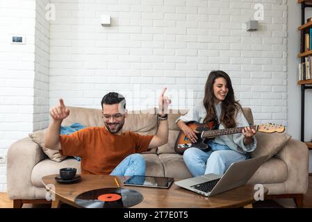 Young creative couple, freelancers web designers taking a break having fun playing electric guitar listening music in their apartment home office list Stock Photo