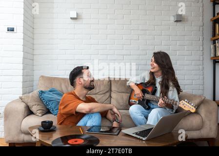 Young creative couple, freelancers web designers taking a break having fun playing electric guitar listening music in their apartment home office list Stock Photo