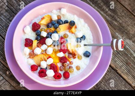 Oatmeal porridge with fruit, berries and mini marshmallow with spoon on wooden background table top view, homemade healthy breakfast cereal Stock Photo