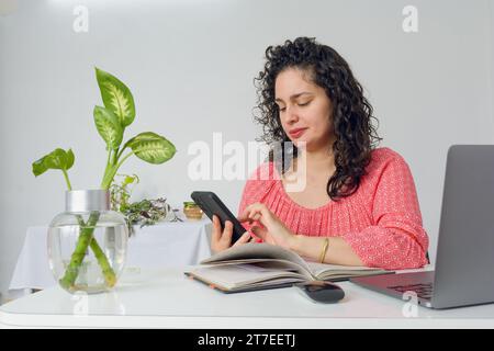 Young latin woman with curlers and business casual clothes working in her home office, sitting using phone sending messages and checking notifications Stock Photo