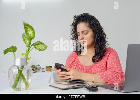 Young latin woman with curlers and business casual clothes working in her home office, sitting using phone sending messages and checking notifications Stock Photo