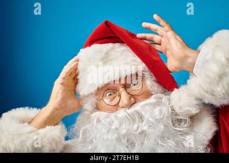portrait of excited Santa Claus with white beard and eyeglasses looking at camera on blue background Stock Photo