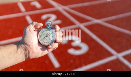Hand holding a stopwatch on a red running track, numbers on lanes visible, sunny day Stock Photo