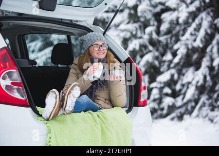 Woman in woolen hat sits in the trunk of the car and holds a cup of hot tea in her hands. Stock Photo