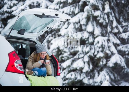 Woman in woolen hat sits in the trunk of the car and holds a cup of hot tea in her hands. Stock Photo