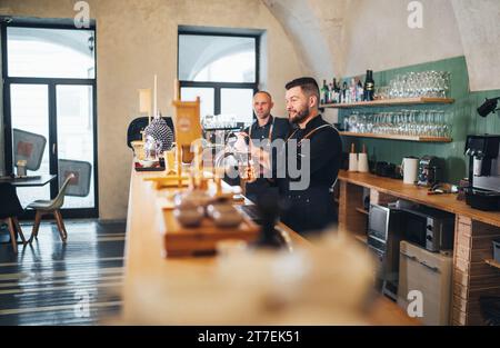 Two smiling caucasian men friends barman lager beer tapping and waiter dressed black uniform at bar counter. People at work, team work, men friendship Stock Photo