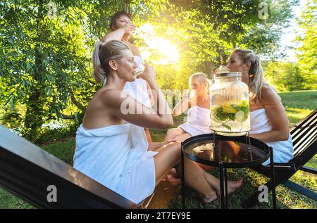 Women in towels drinking cold lemonade with lemon slices outdoors after sauna at spa resort Stock Photo