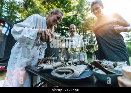 woman pours white wine for friends at an outdoor table set with plates and napkins, after a finnish sauna day Stock Photo