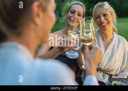 Happy women in bathrobes toasting with white wine glasses outdoors, enjoying a spa day Stock Photo