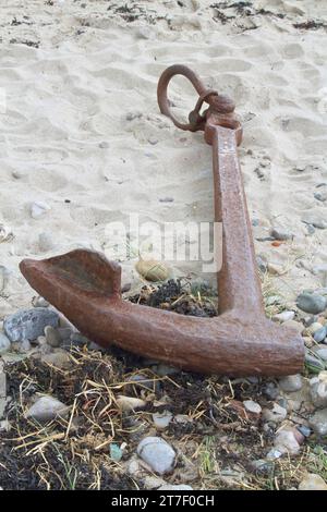 old rusty beached anchor on the sand with seaweed and rocks close by Stock Photo