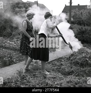 1960s, historical, two ladies burning vegetation on a bonfire in a back garden, one lady in a sleeveless top and pleated skirt, using a garden fork to add leaves to the fire, England, Stock Photo