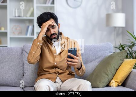 Sad unhappy and disappointed man sitting at home on sofa in living room, Indian man received notification message with bad news, reading using smartphone app. Stock Photo