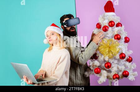 Christmas family using VR glasses and laptop. Bearded man in virtual reality goggles headset. Woman in Santa hat with notebook. Christmas couple near Stock Photo