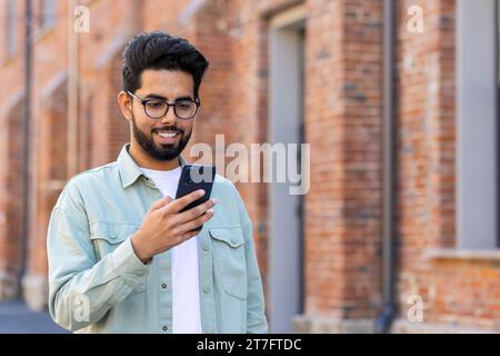 A young man walks through the city with a phone in his hands, a businessman in casual clothes uses an application on a smartphone, smiles, reads online pages, types a text message. Stock Photo