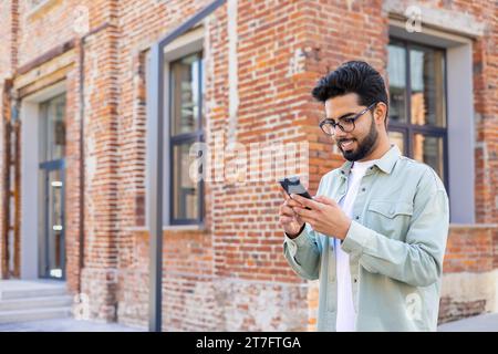 A young man walks through the city with a phone in his hands, a businessman in casual clothes uses an application on a smartphone, smiles, reads online pages, types a text message. Stock Photo
