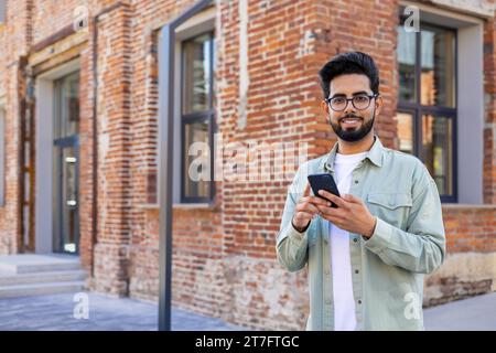 Portrait of young successful student outside university campus, man smiling looking at camera and using app on phone, typing message and browsing social media, Indian man holding phone with backpack. Stock Photo