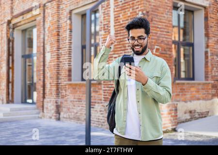 Young indian student received online win notification message phone, successful entrance exam results, man with backpack walking outside university campus, celebrating successful achievement result. Stock Photo