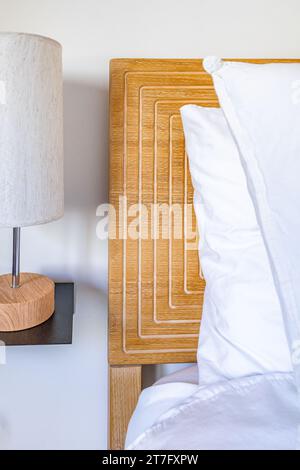 Bedroom with bright wooden headboard, white linens, and nightstand table with lamp Stock Photo