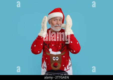 Senior man in Christmas sweater, Santa hat and knitted mittens on light blue background Stock Photo