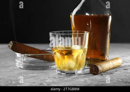 Bottle, glass of whiskey with ice cubes and cigars on grey table Stock Photo