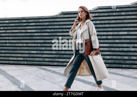 Stylish woman in a coat walking outdoors talking on a mobile phone. Businesswoman with ginger hair and a leather folder walking on the street. Stock Photo