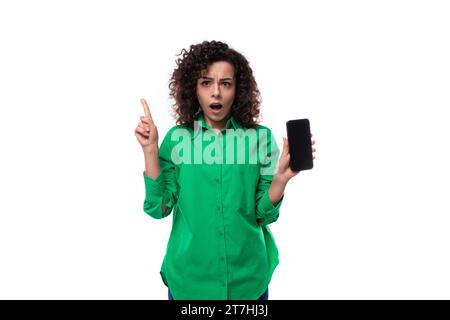 young employee of the company woman with black hair dressed in a green shirt shows the smartphone screen in surprise Stock Photo