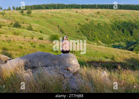 Young adult blonde woman sitting on rock with cellphone at summer day in wilderness Stock Photo