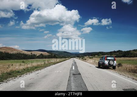 roadside car on rural road trip landscape below white clouds in blue sky of Bosnia and Herzegovina Stock Photo