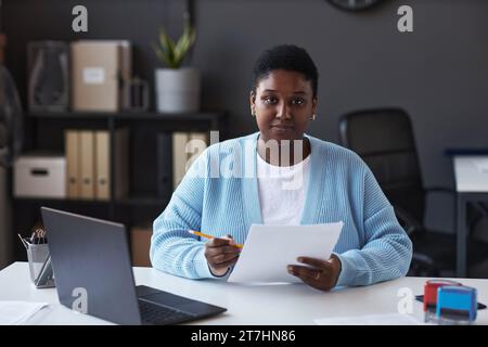Young confident female manager of visa application center in casualwear looking at camera while sitting by workplace and checking data Stock Photo