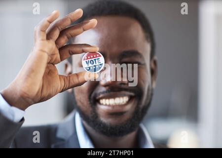 Focus on hand of young smiling African American man holding vote badge in front of his right eye while standing in front of camera Stock Photo