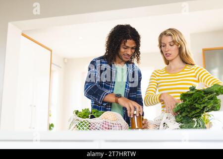 Happy diverse couple unpacking grocery shopping and discussing in sunny kitchen Stock Photo