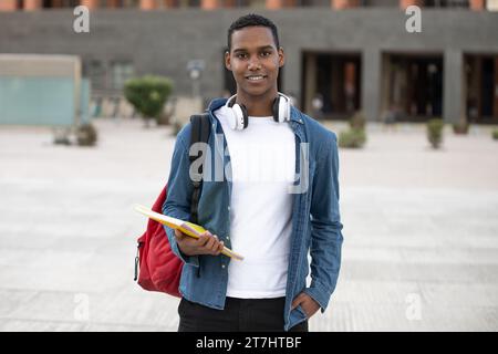 Handsome student wearing a backpack and holding a notebook looking at camera.Relaxed and happy man standing outside with headphones ready for college. Stock Photo