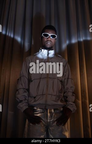 Dramatic fashion portrait of young Black man wearing sunglasses and looking at camera posing in silver outfit Stock Photo