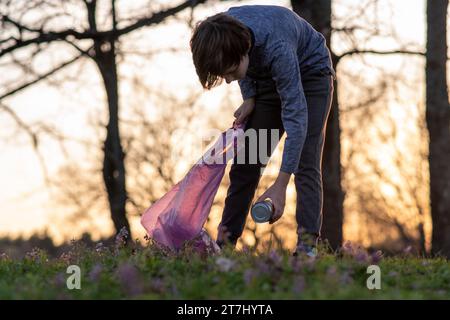 Teenage boy collects garbage in big pink bag in nature, on meadow on autumn evening at sunset. Environmental problems. We take care of nature. Saving Stock Photo