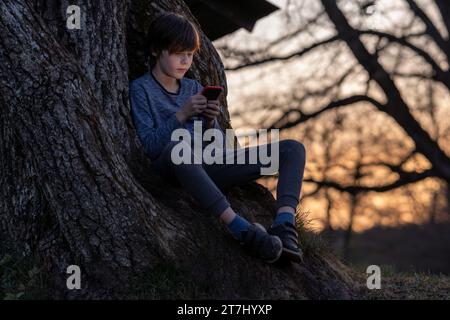 Portrait of teenage boy sitting on roots of large tree and enthusiastically watching something on his phone against background of setting sun. Summer Stock Photo