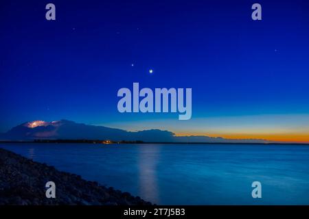 A scene of the crescent Moon with Earthshine, planets and the last of the winter stars in the evening twilight, with a thunderstorm and lightning bolt Stock Photo