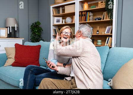 Happy senior old couple holding smartphone looking at cellphone screen laughing relaxing sit on sofa together, smiling elder mature grandparents famil Stock Photo