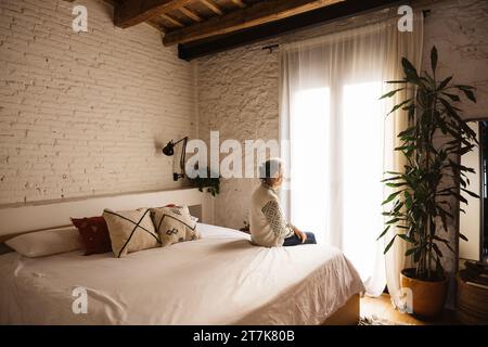 Portrait of thoughtful real mature senior woman sitting on bed in front of a window in a bedroom. Stock Photo