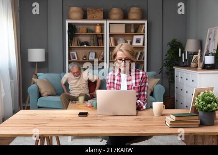 Senior woman sitting at desk with laptop computer watching online educational course. Older retired female video call with her friend while husband si Stock Photo