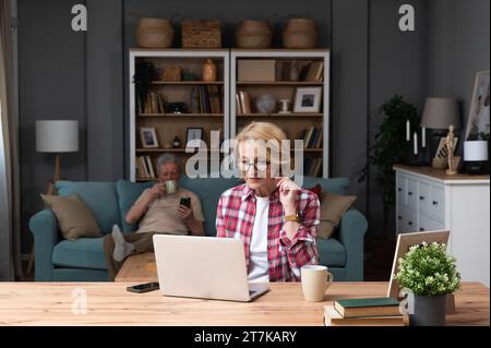 Senior woman sitting at desk with laptop computer watching online educational course. Older retired female video call with her friend while husband si Stock Photo