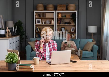 Senior woman sitting at desk with laptop computer watching online educational course. Older retired female video call with her friend while husband si Stock Photo