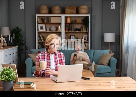 Senior woman sitting at desk with laptop computer watching online educational course. Older retired female video call with her friend while husband si Stock Photo