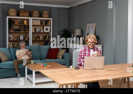 Senior woman sitting at desk with laptop computer watching online educational course. Older retired female video call with her friend while husband si Stock Photo