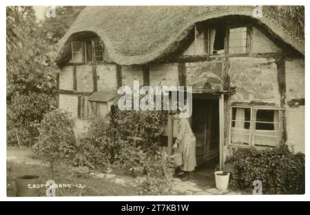 Original early1900's postcard of a lady with pail / bucket outside her charming typical English half timbered thatched cottage, vernacular architecture, dating from Medieval period, Cropthorne, Worcestershire. England, U.K. Circa 1917 Stock Photo