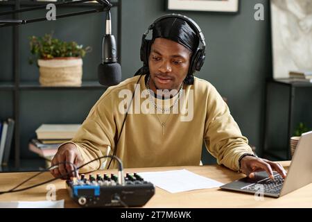 African American guy in headphones switching consoles on soundboard and adjusting sound before recording new podcast in home studio Stock Photo