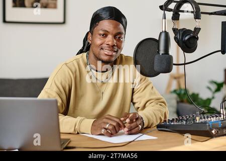 Young smiling man in stylish casualwear sitting by workplace equipped with microphone, headphones, soundboard and laptop Stock Photo
