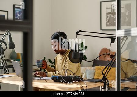 Young African American man with cup of coffee using laptop at break while preparing for recording new podcast with guest in studio Stock Photo