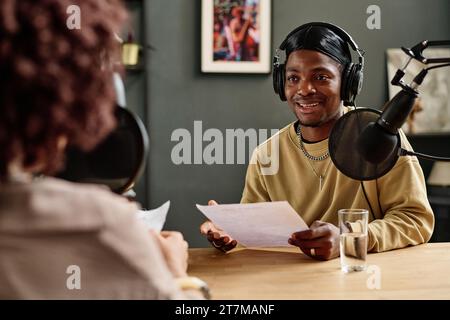 Young African American man with paper speaking in microphone while explaining something or asking question to guest invited for interview Stock Photo